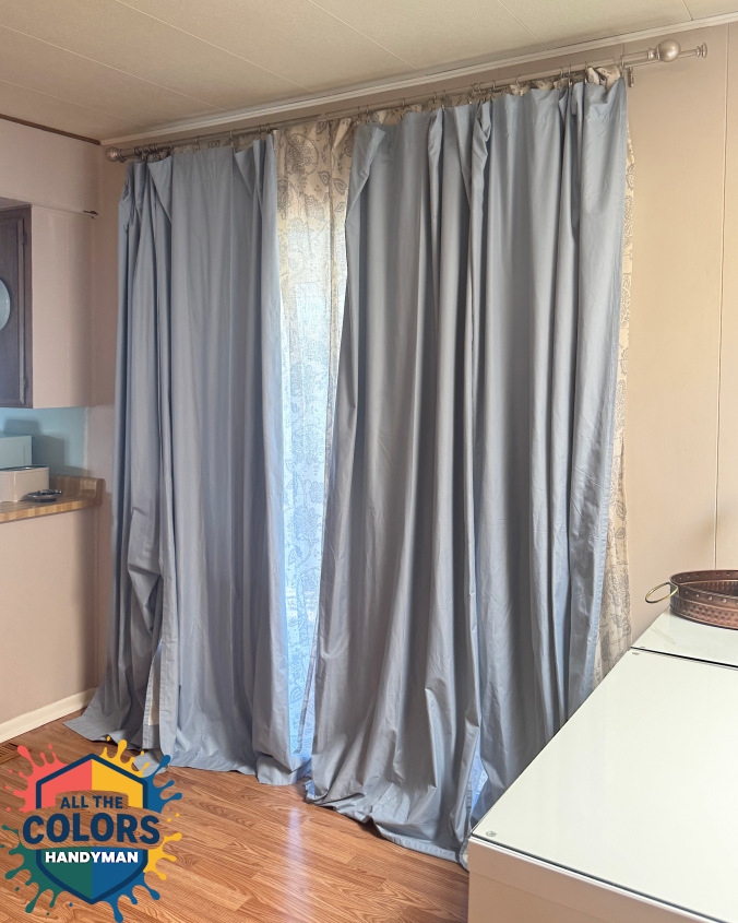 Interior photo of a patio door with curtains in front of it. A sheer grey-patterned curtain hangs closest to the door. It's mostly covered by a light blue blackout curtain. Both curtains are pooling on the hardwood floor.